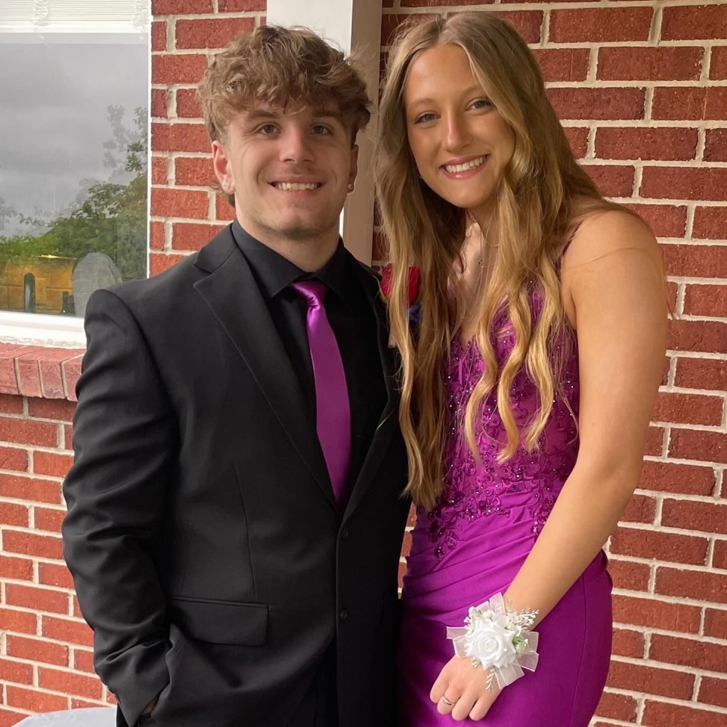 Man in a black suit with pink tie, woman in a pink gown, smiling; posing in front of brick wall.