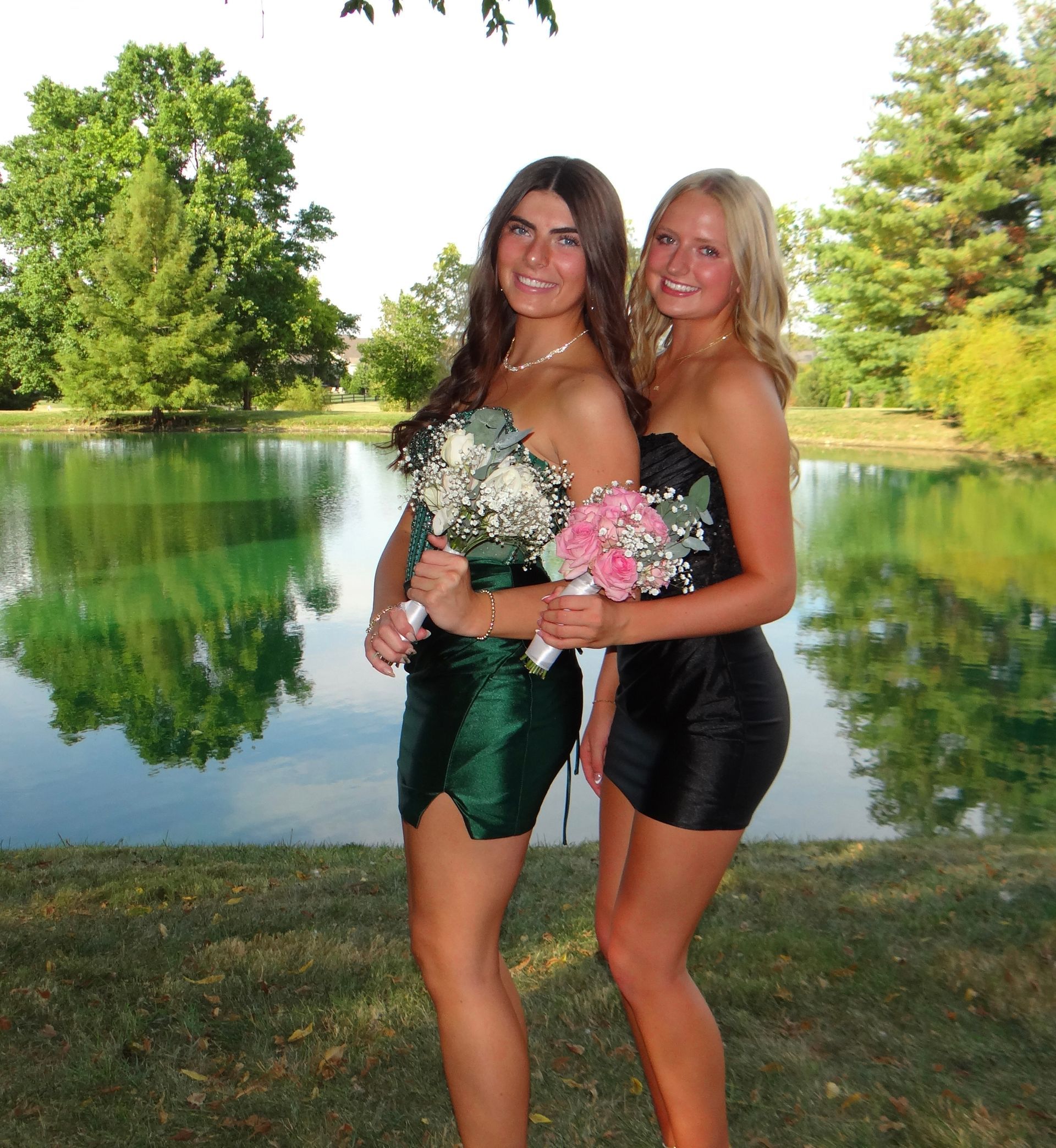 Two women in short formal dresses, holding bouquets, pose by a lake.