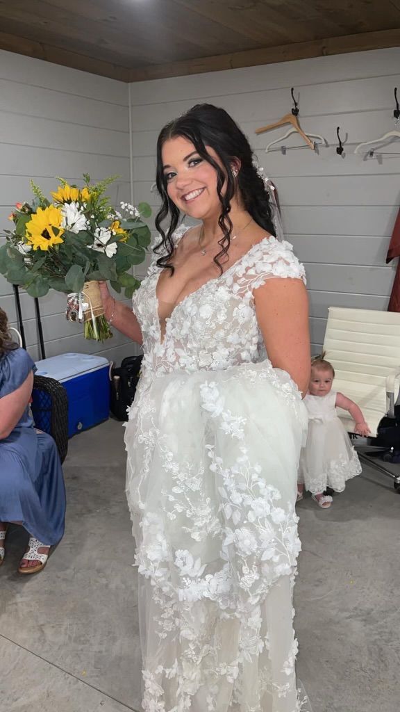Bride in a white floral wedding dress holding a bouquet of sunflowers and greenery, smiling.