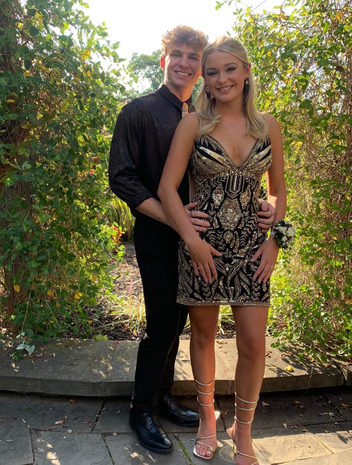 Couple in formal attire; woman in beaded black and gold dress, man in black shirt and pants, outside by greenery.