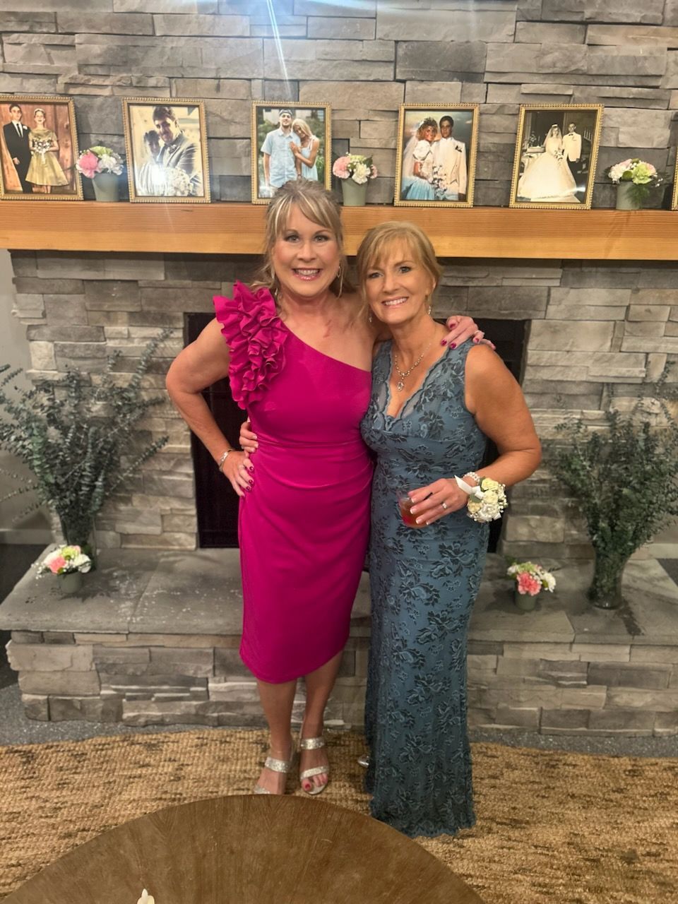 Two women pose in front of a stone fireplace, one in a pink, one-shoulder dress, the other in a blue gown.