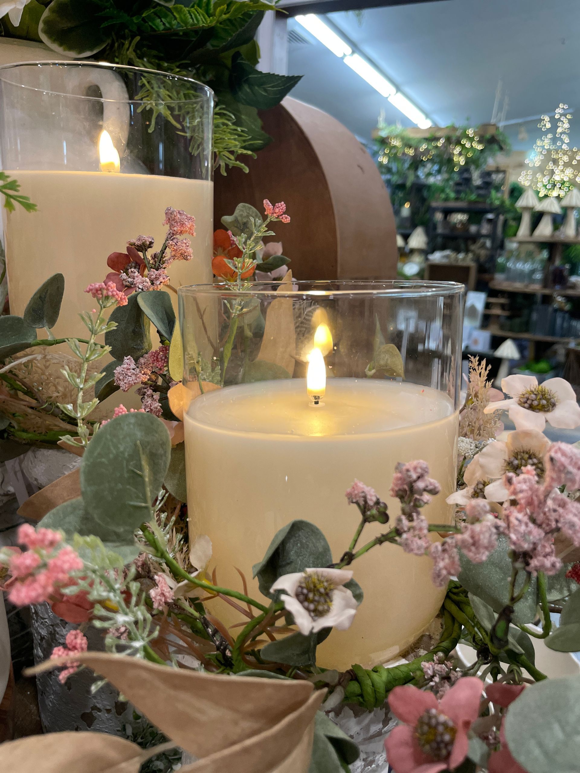 Two lit cream-colored pillar candles in glass holders, surrounded by flowers and greenery in a store.