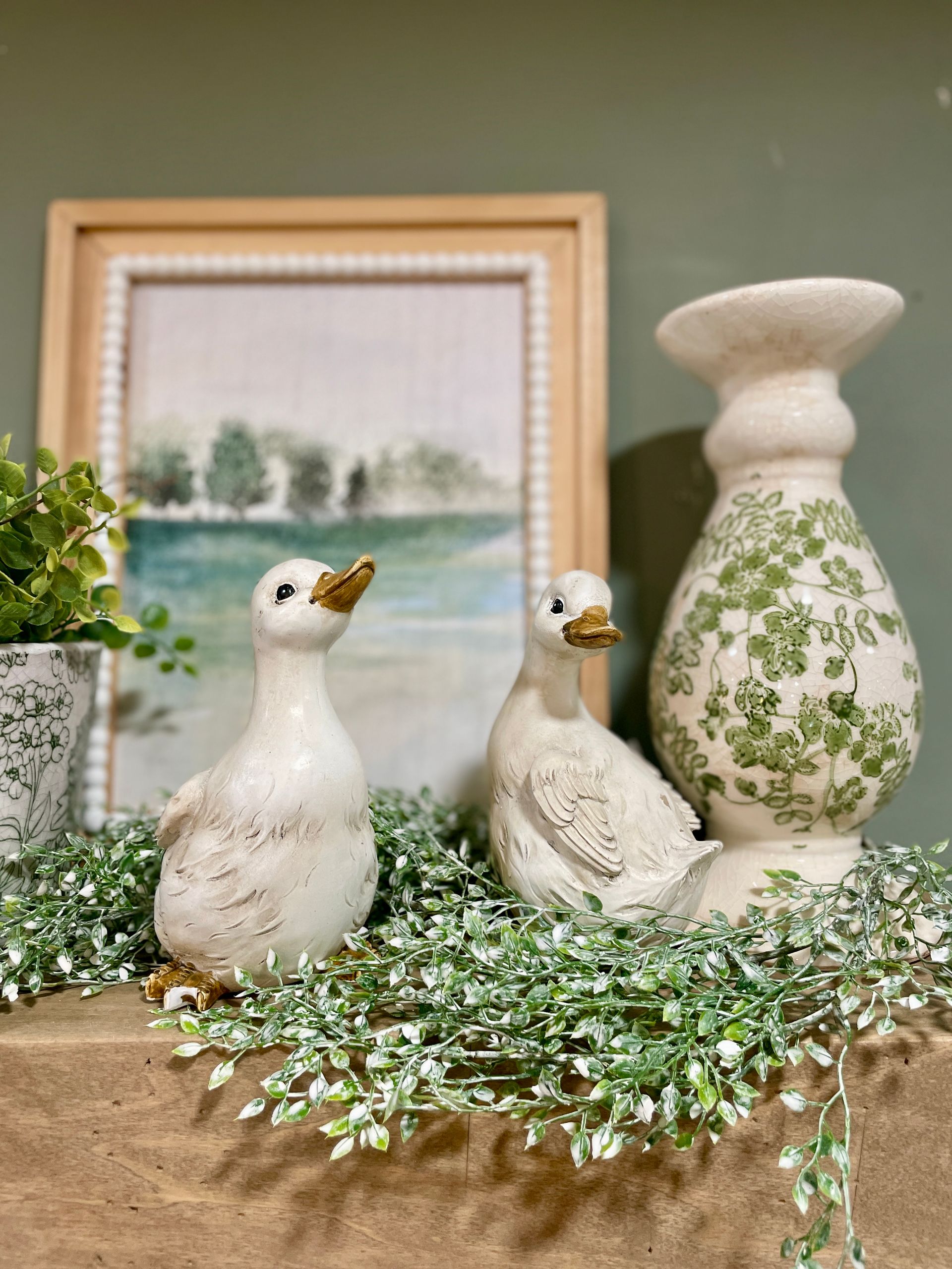 Two ceramic duck figurines sit on a shelf with greenery and a framed landscape painting.