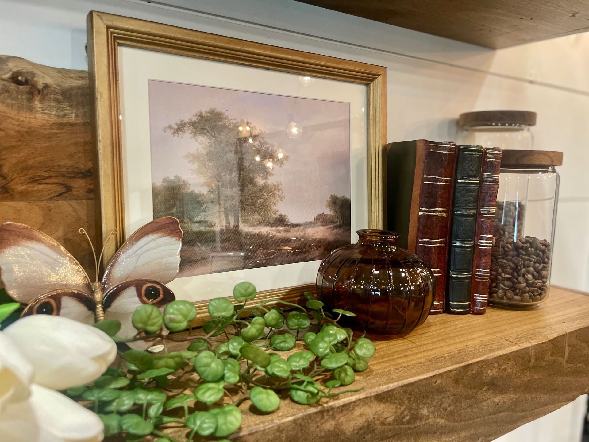 Shelf decor: framed landscape art, antique books, glass jars with coffee beans, faux greenery, and a butterfly.