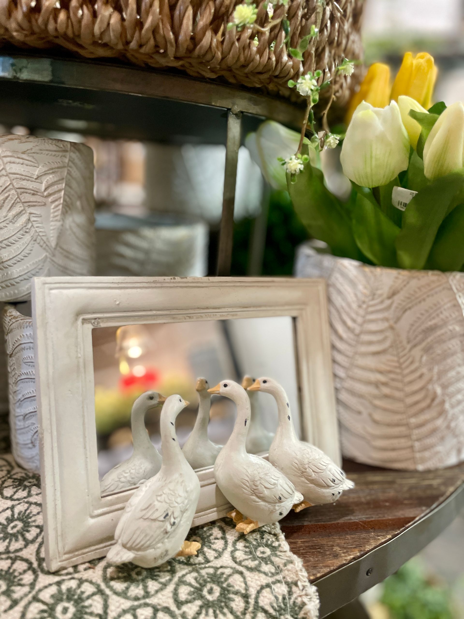 Three goose figurines near a mirror, with tulips and decorative objects on a shelf.