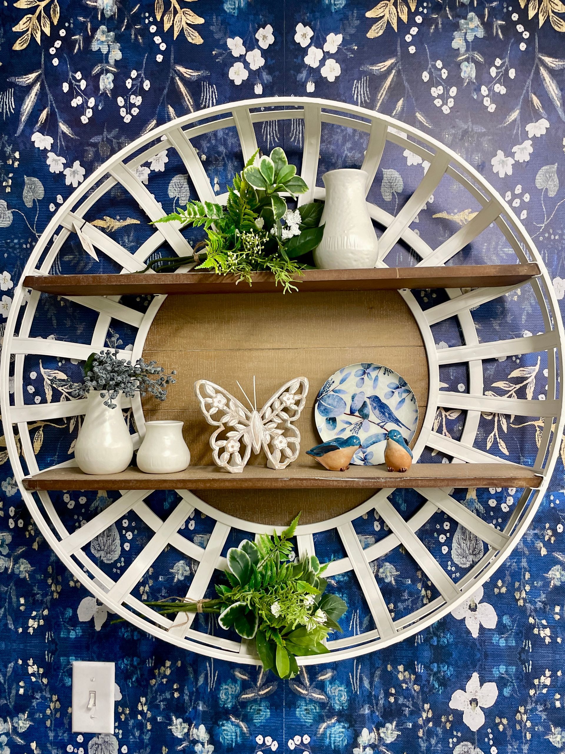 White, circular wall shelf with two wooden shelves, displaying decor against a blue floral wallpaper.