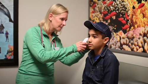 A healthcare professional in a green top uses a digital thermometer on a patient's forehead in a room with wall art.
