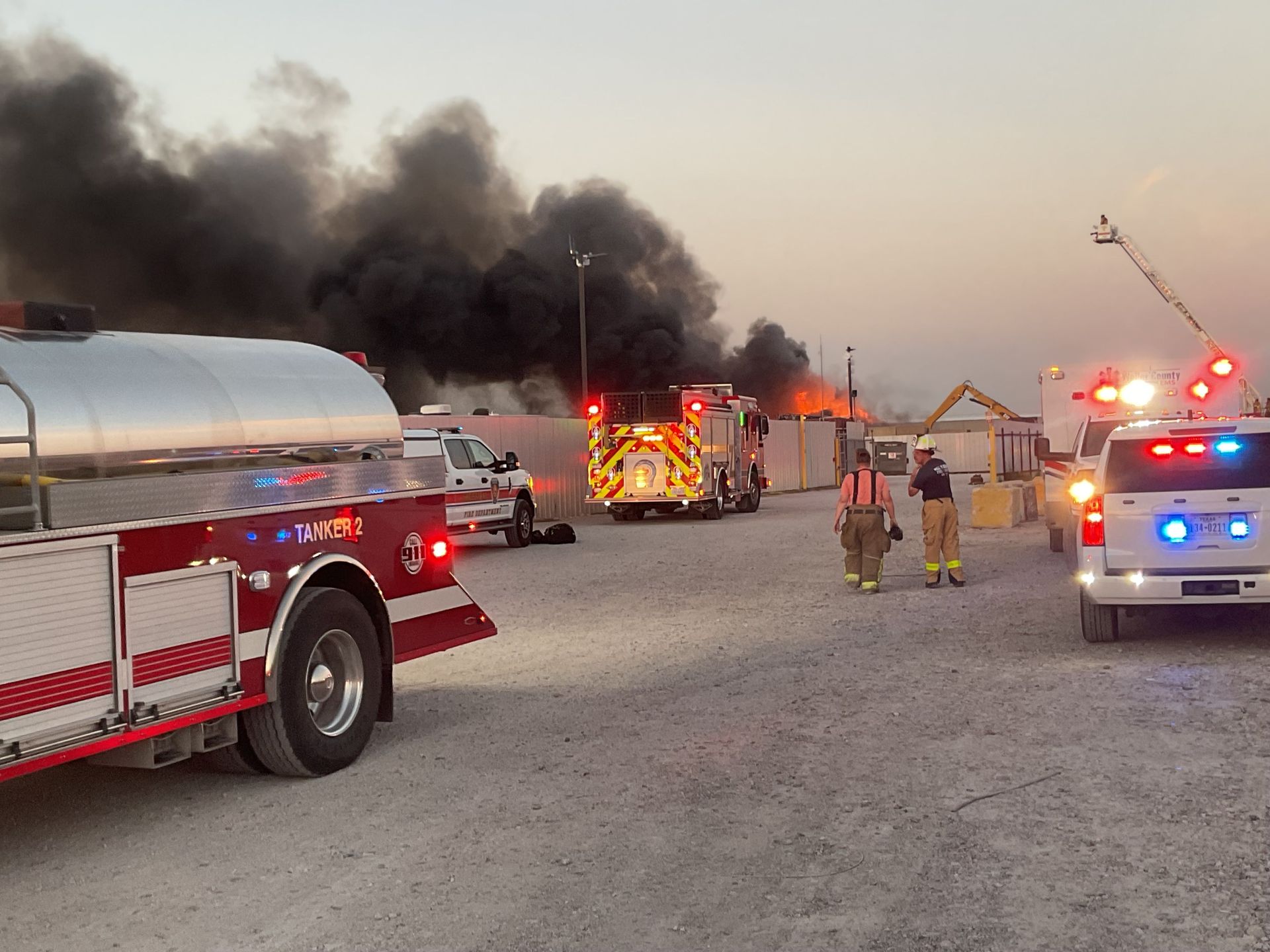 A group of fire trucks are parked in a gravel lot with an aerial fire truck spraying water onto a fire.