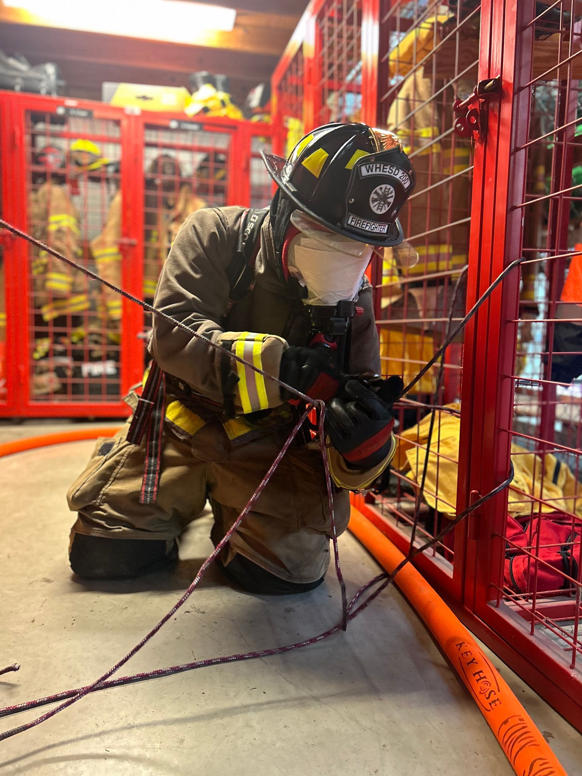 A firefighter is kneeling down in front of a red cage holding a rope.