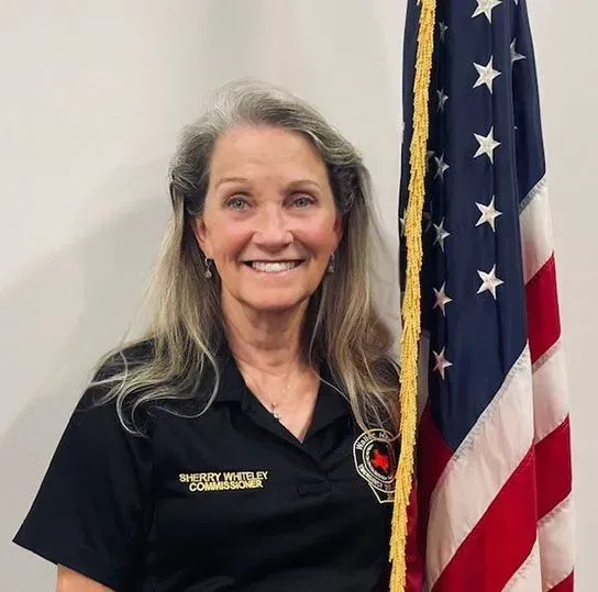 A picture of Commissioner Sherry Whiteley wearing a black shirt standing next to an American Flag.