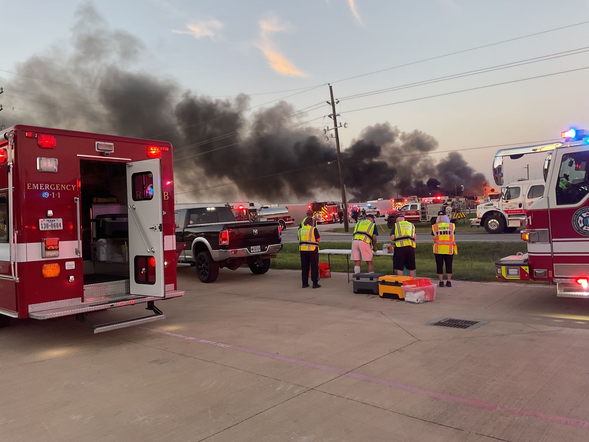 A group of fire trucks are parked in a parking lot in front of a large fire.