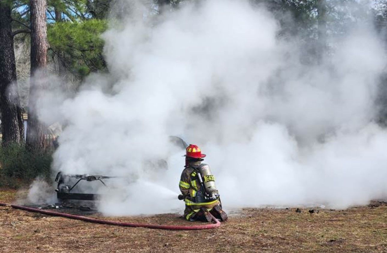 a fire fighter is kneeling while spraying water on a car fire.