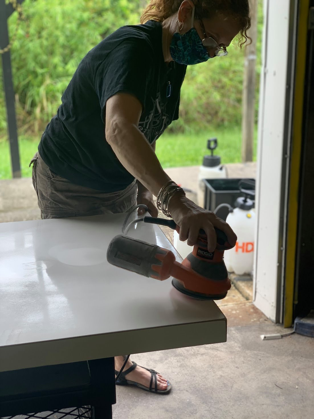 A woman wearing a mask is sanding a table with a sander.
