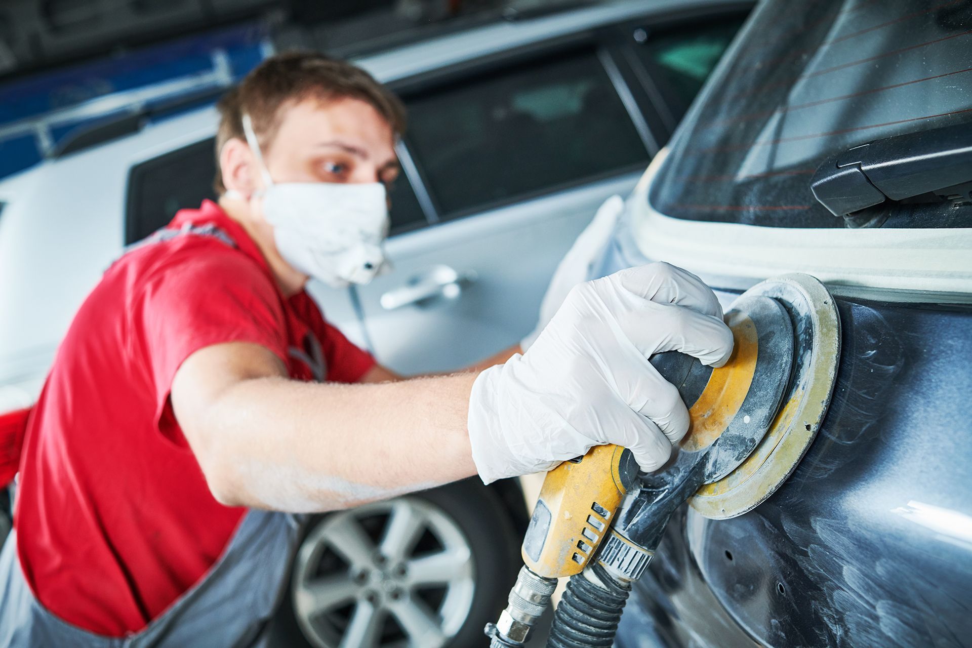 A man wearing a mask and gloves is sanding a car.
