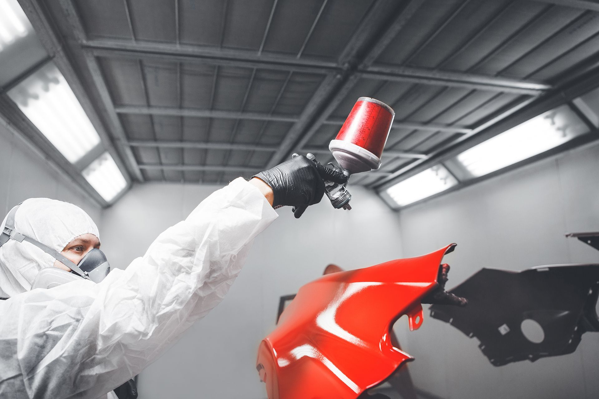 A man is painting a red car bumper in a paint booth.