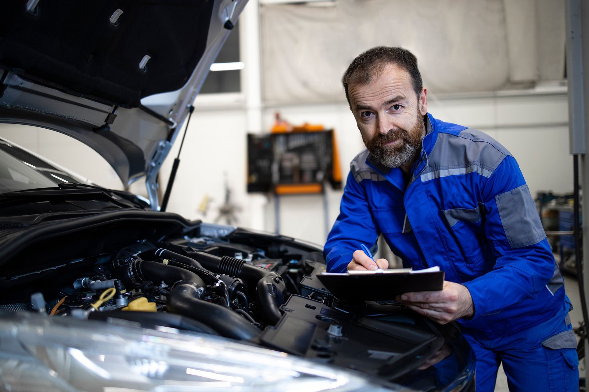 A mechanic is looking under the hood of a car while holding a clipboard.