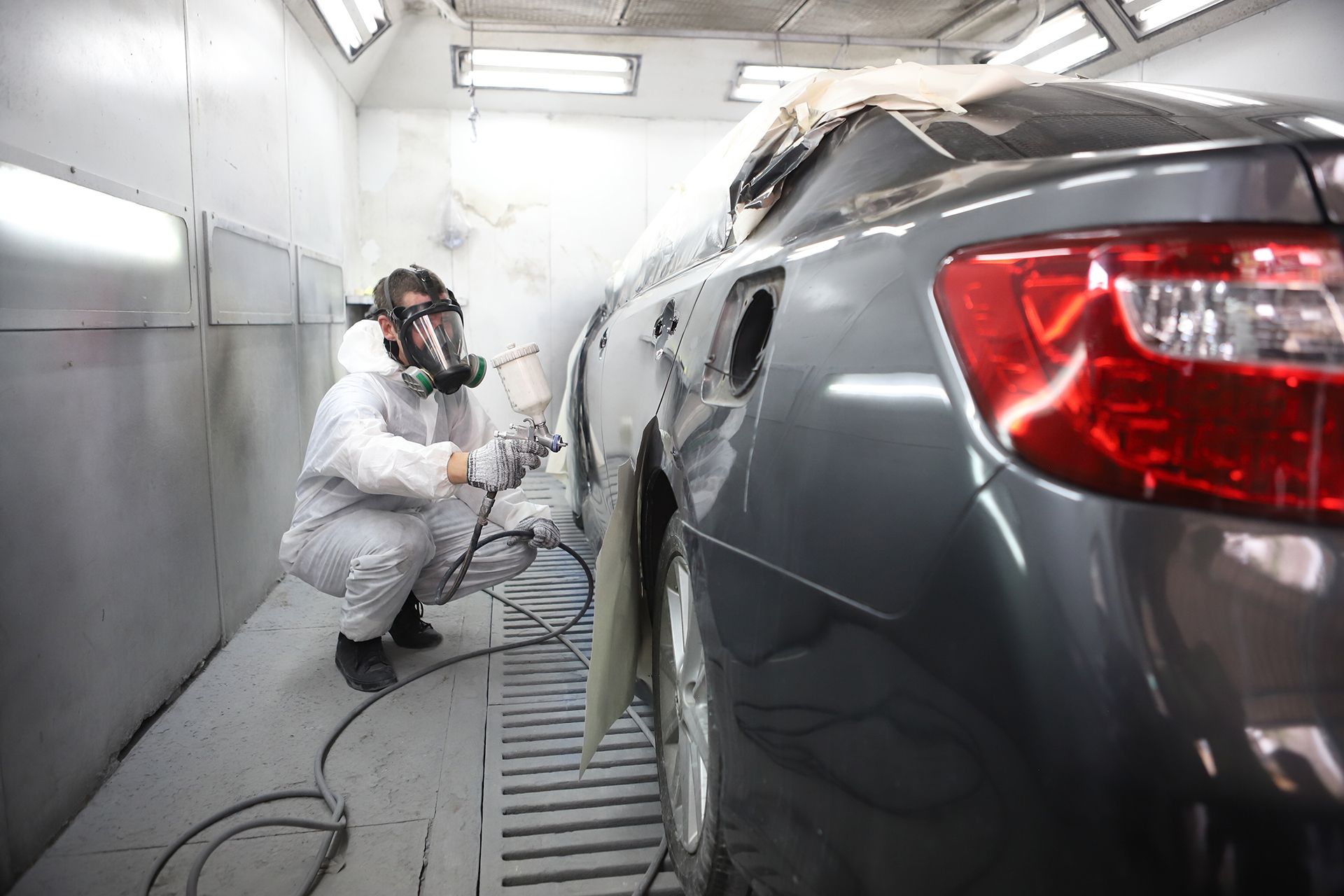 A man wearing a gas mask is painting a car in a paint booth.