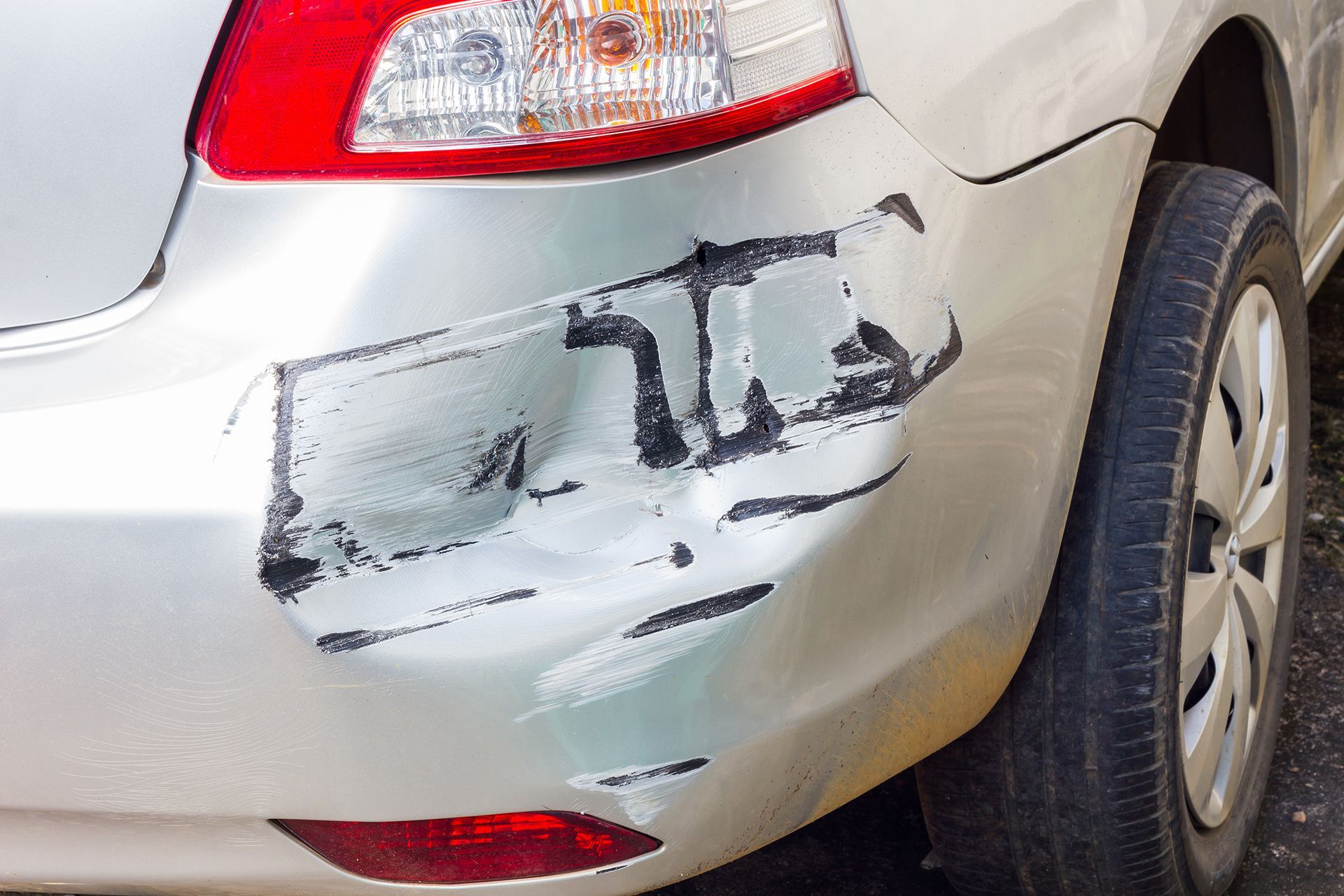 A silver car with a damaged bumper is parked on the side of the road.