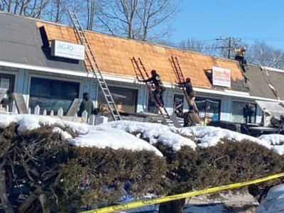 Roofers replacing a roof on a building covered in snow, with ladders and safety tape present.