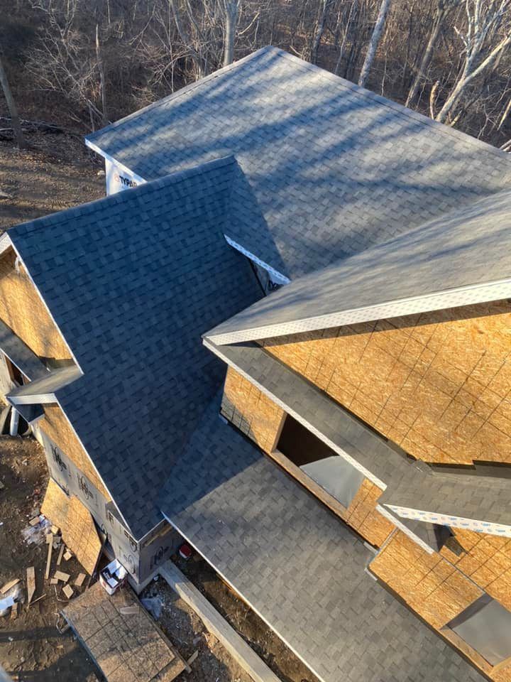Partially constructed house with dark gray shingled roofs and exposed wooden framing. Sunlight reflects on the surfaces