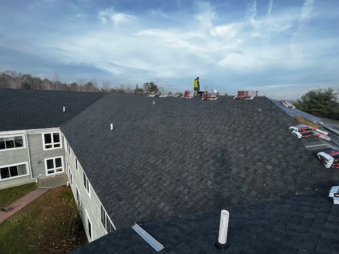 Workers installing shingles on a dark gray roof of a multi-story building under a cloudy sky.