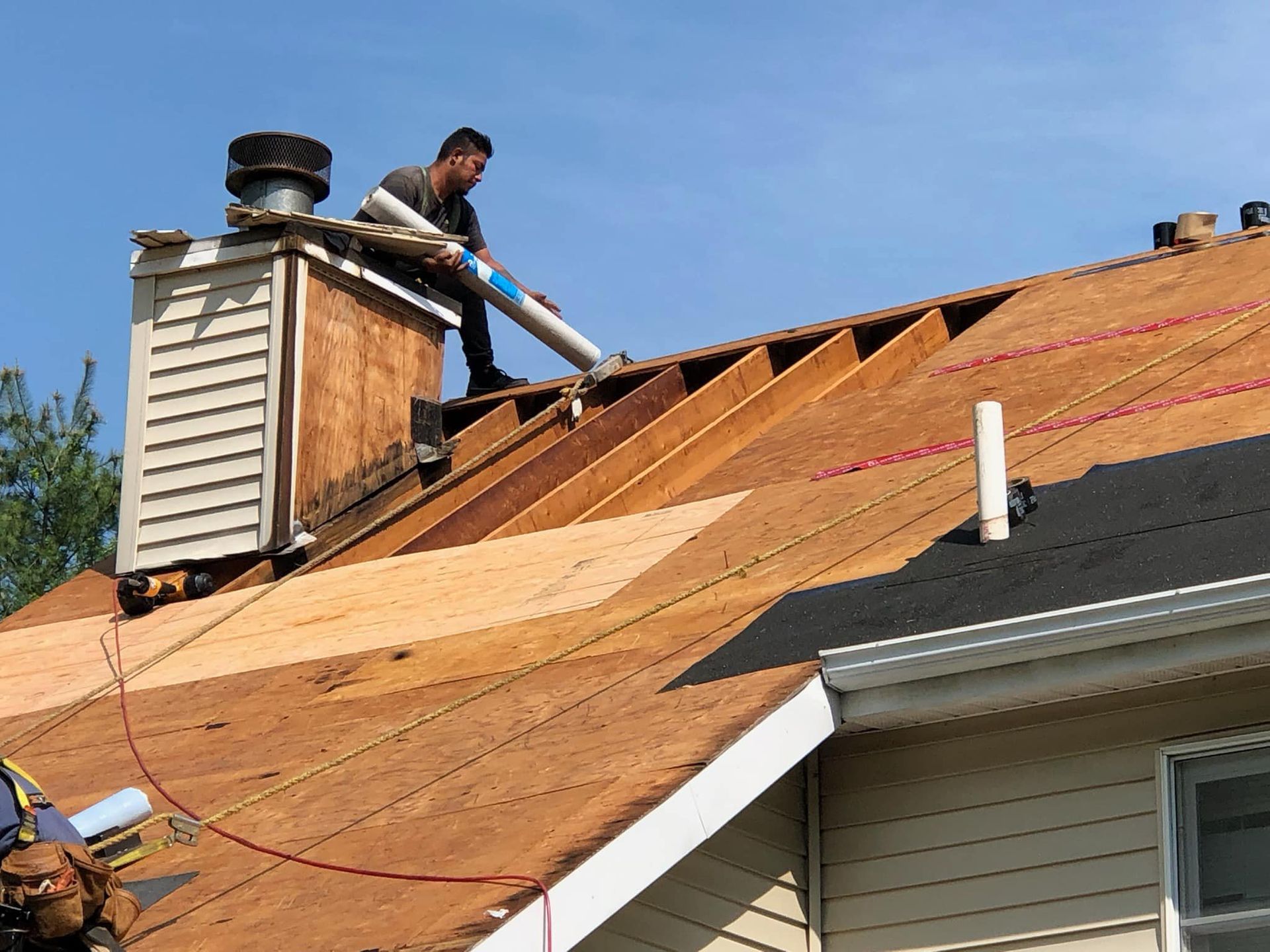 A construction worker on a roof applies adhesive to a pipe. The sky is blue, and the roof is partially covered with plywood.