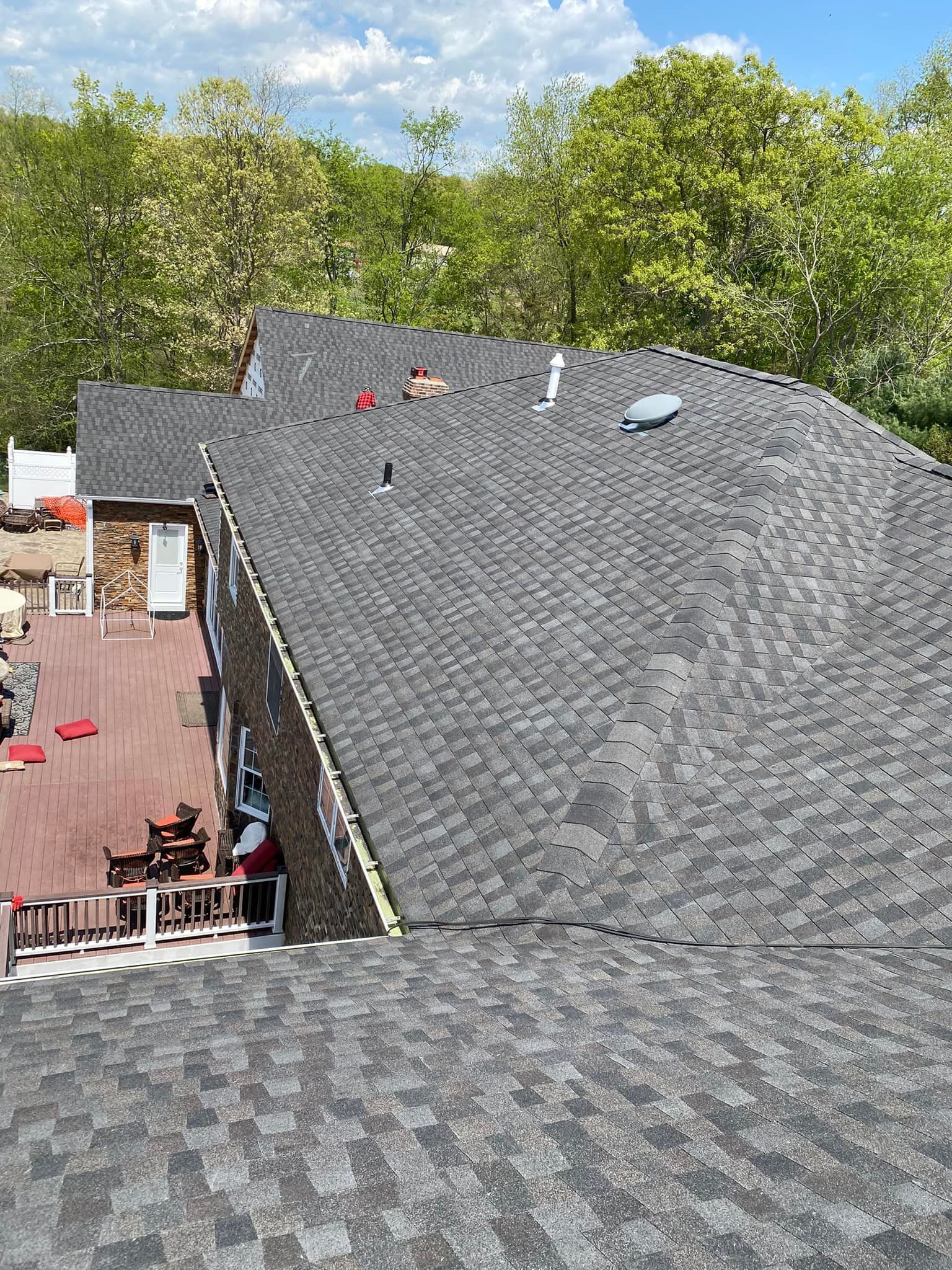 View of a house with a dark gray shingled roof, a deck, and trees in the background under a sunny sky.
