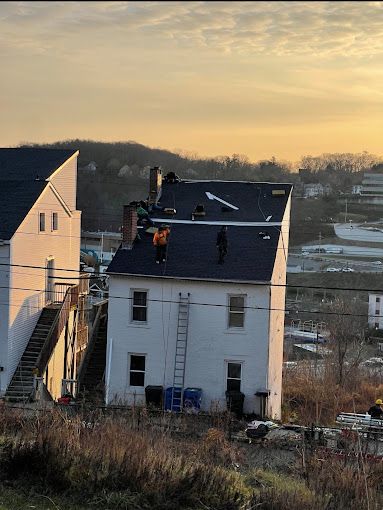 Two workers on a white house roof in an orange sunset. A ladder leans against the building.