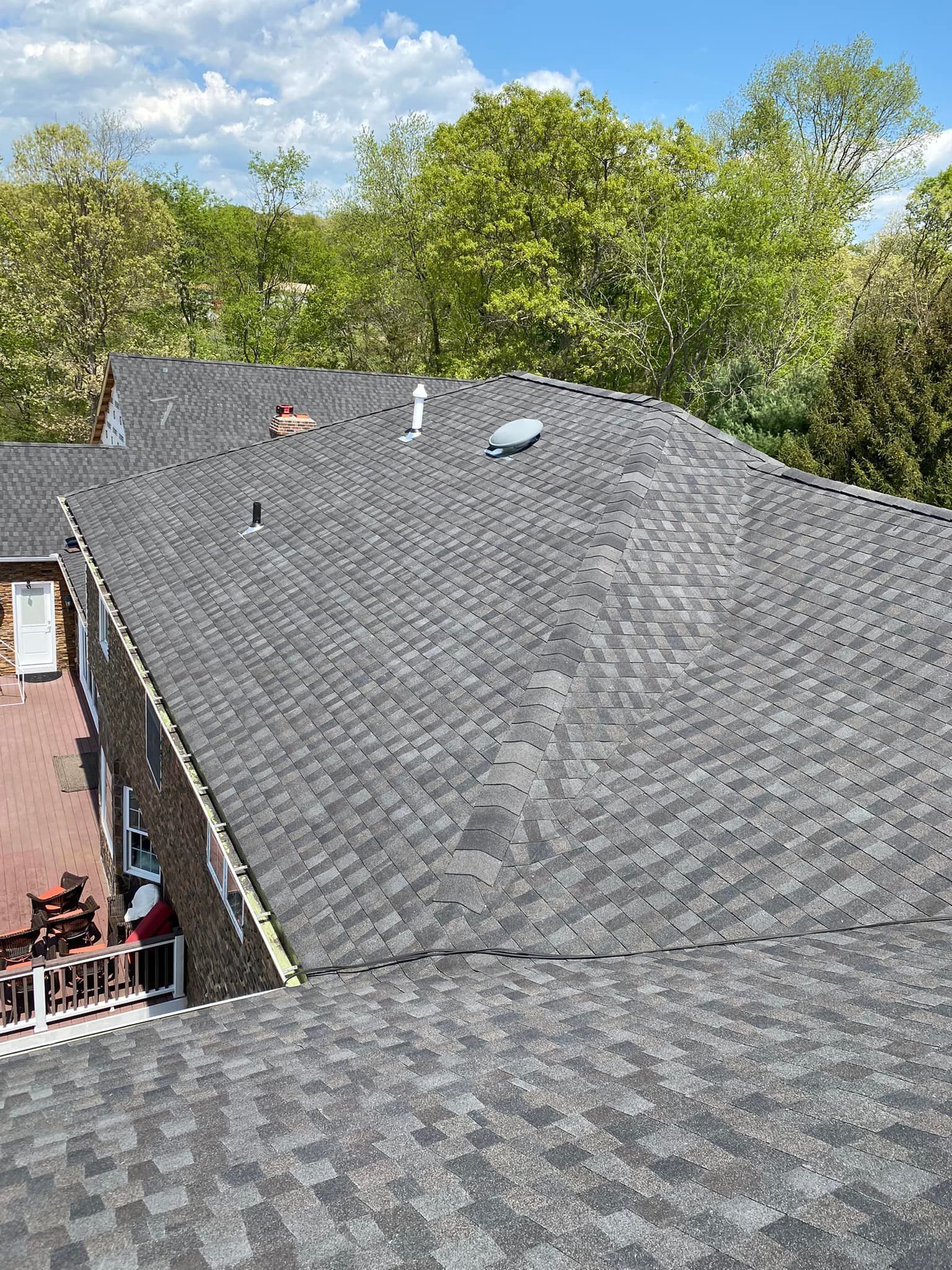Dark gray asphalt shingle roof with multiple vents. Deck visible on the left, trees and blue sky in the background.