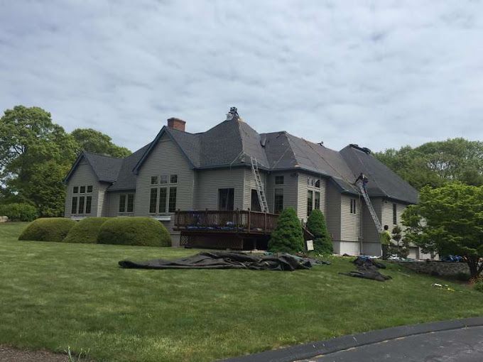 A large house with dark gray roofing being worked on. Green grass and trees surround it.
