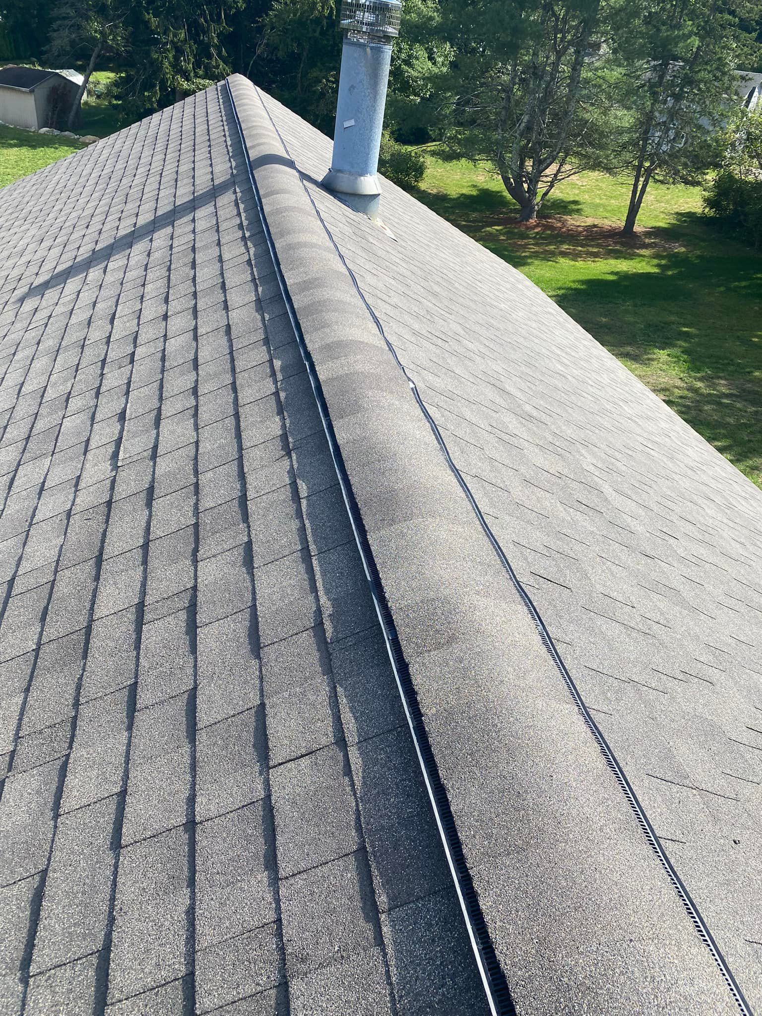 Close-up view of a gray asphalt shingle roof with a chimney and ridge cap, set outdoors on a sunny day.