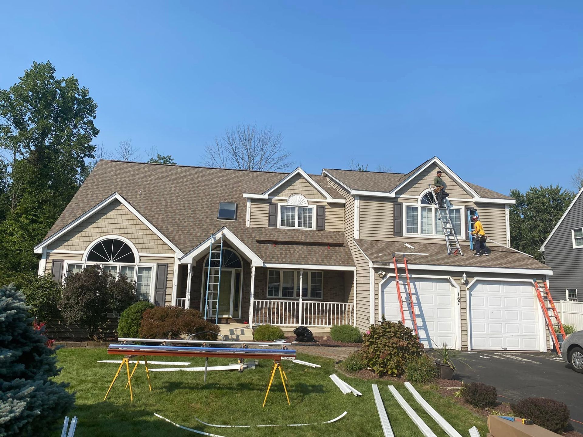 Two-story beige house with roof repair in progress; workers on the roof with ladders; green lawn and blue sky.