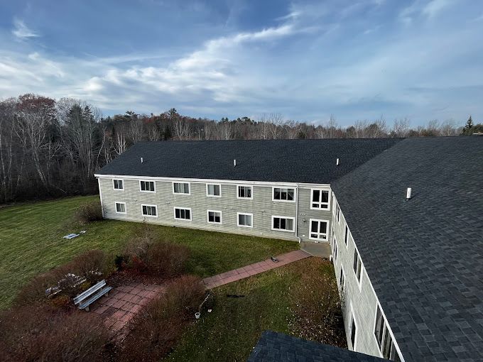 Two-story gray building with a dark roof. A red brick patio and grass lawn are in the foreground, with a forest