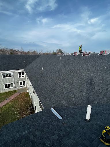 Roofer in a safety vest working on a dark asphalt shingle roof on a sunny day.