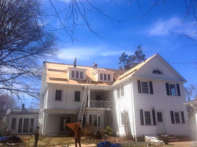 Workers on a white house roof installing shingles. The blue sky is clear, with some trees visible.