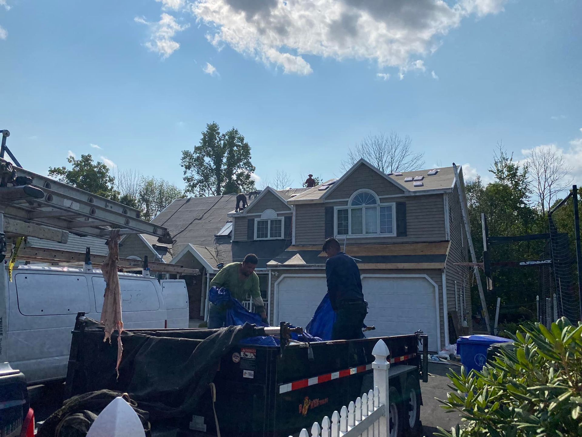 Roofers working on a house; two men in a truck, with dumpsters, ladders, and the house under construction on a sunny day.