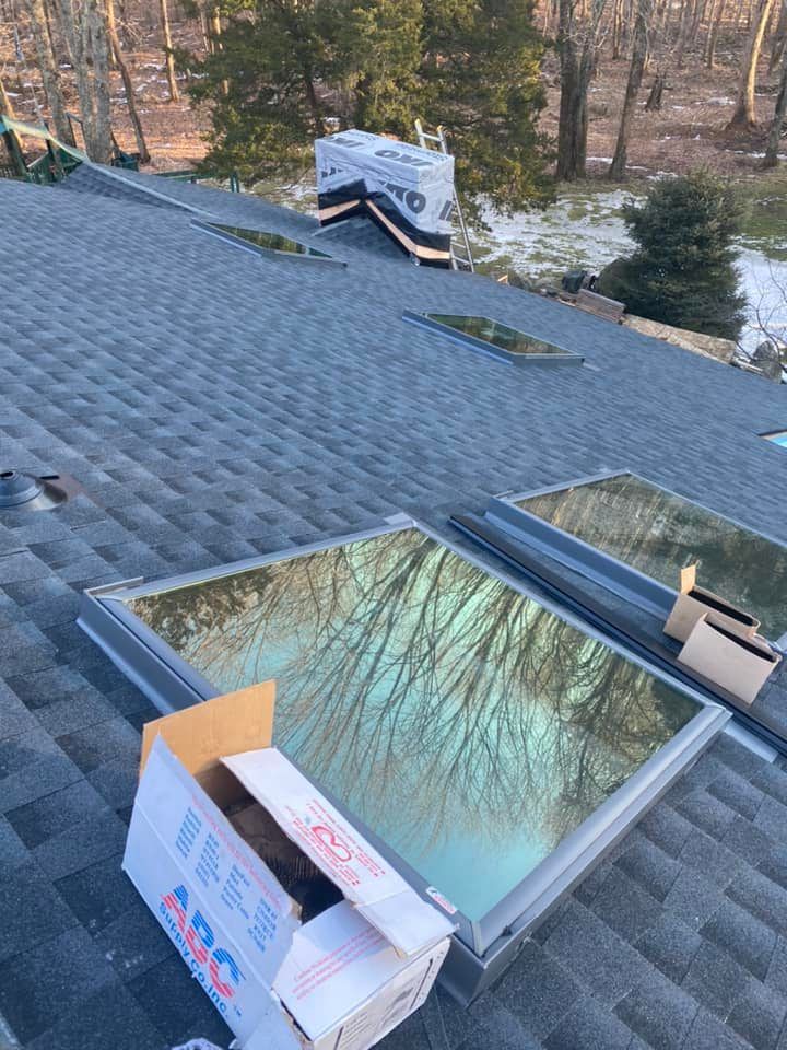 A roof with installed skylights reflects trees and sky. Cardboard boxes and roofing materials are present.
