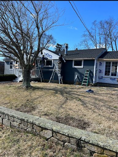 Construction workers on ladders installing siding on a blue house with a gray roof; a sunny day.