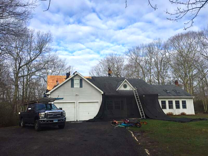 A house undergoing roofing repairs; workers are on the roof with a truck, ladder, and tarp on the driveway.