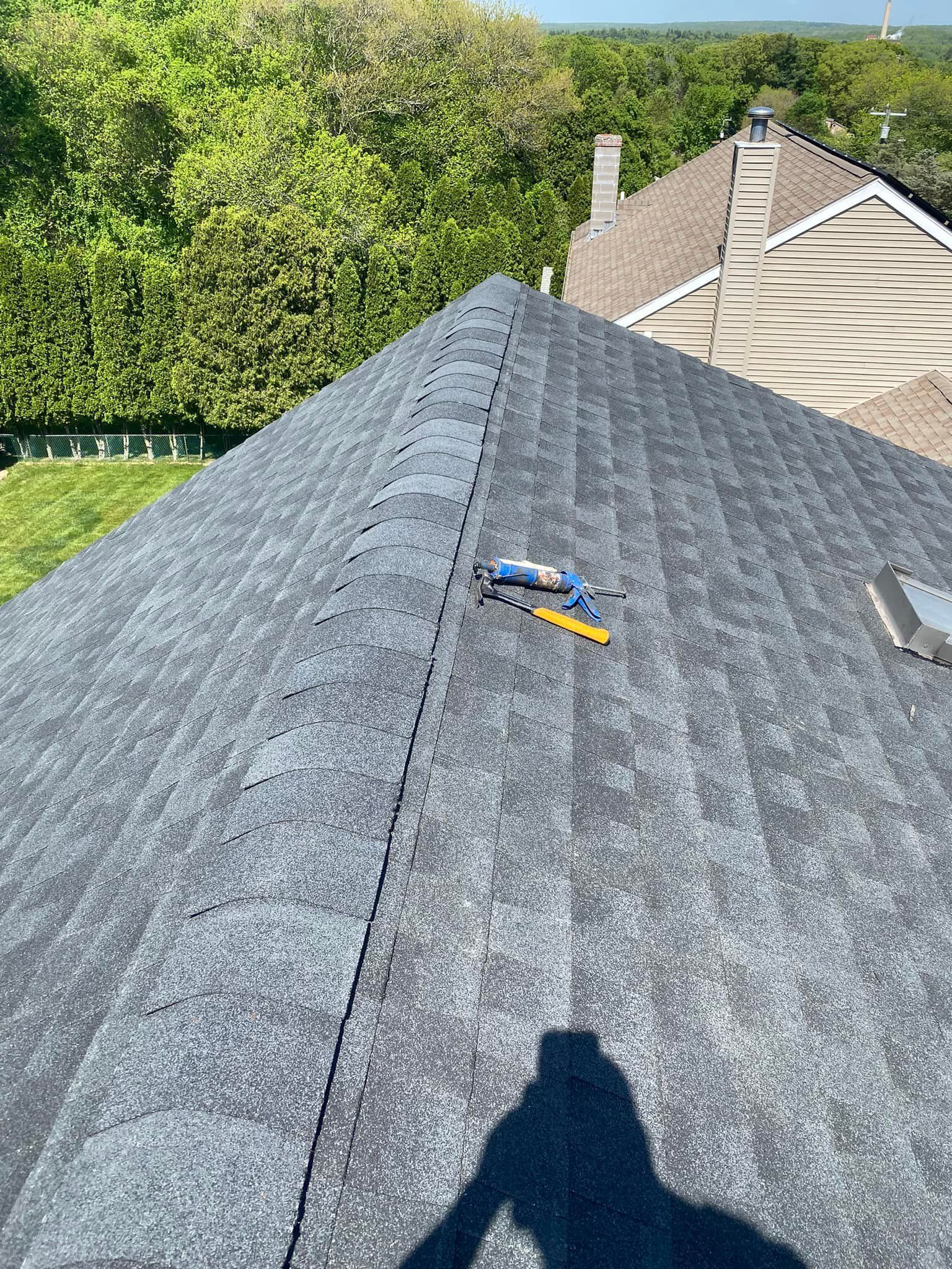 Looking down at a dark shingled roof, tools visible near the ridge, with a leafy green background.