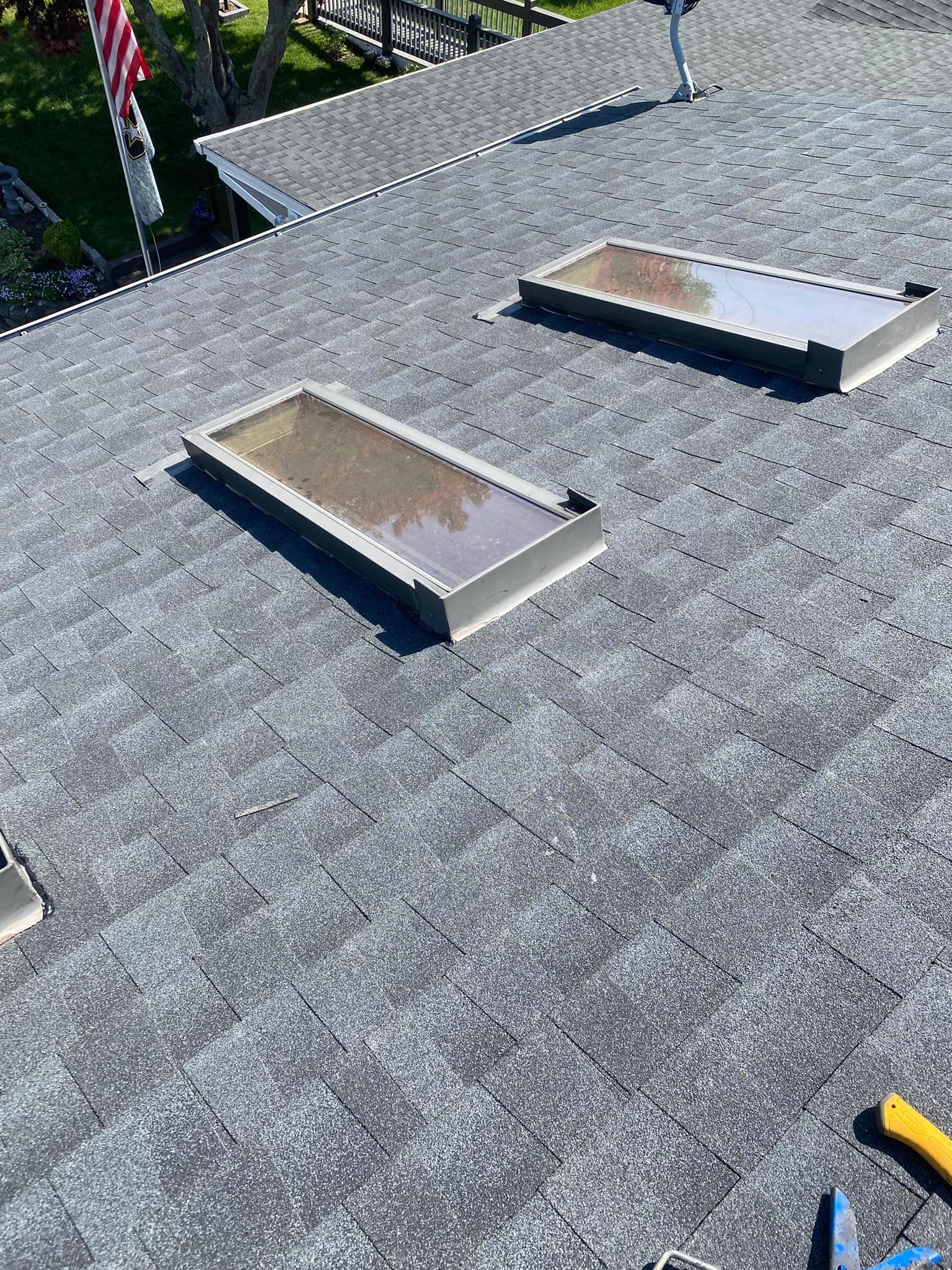 View from above of a gray shingled roof with two rectangular skylights. A US flag flies in the distance.