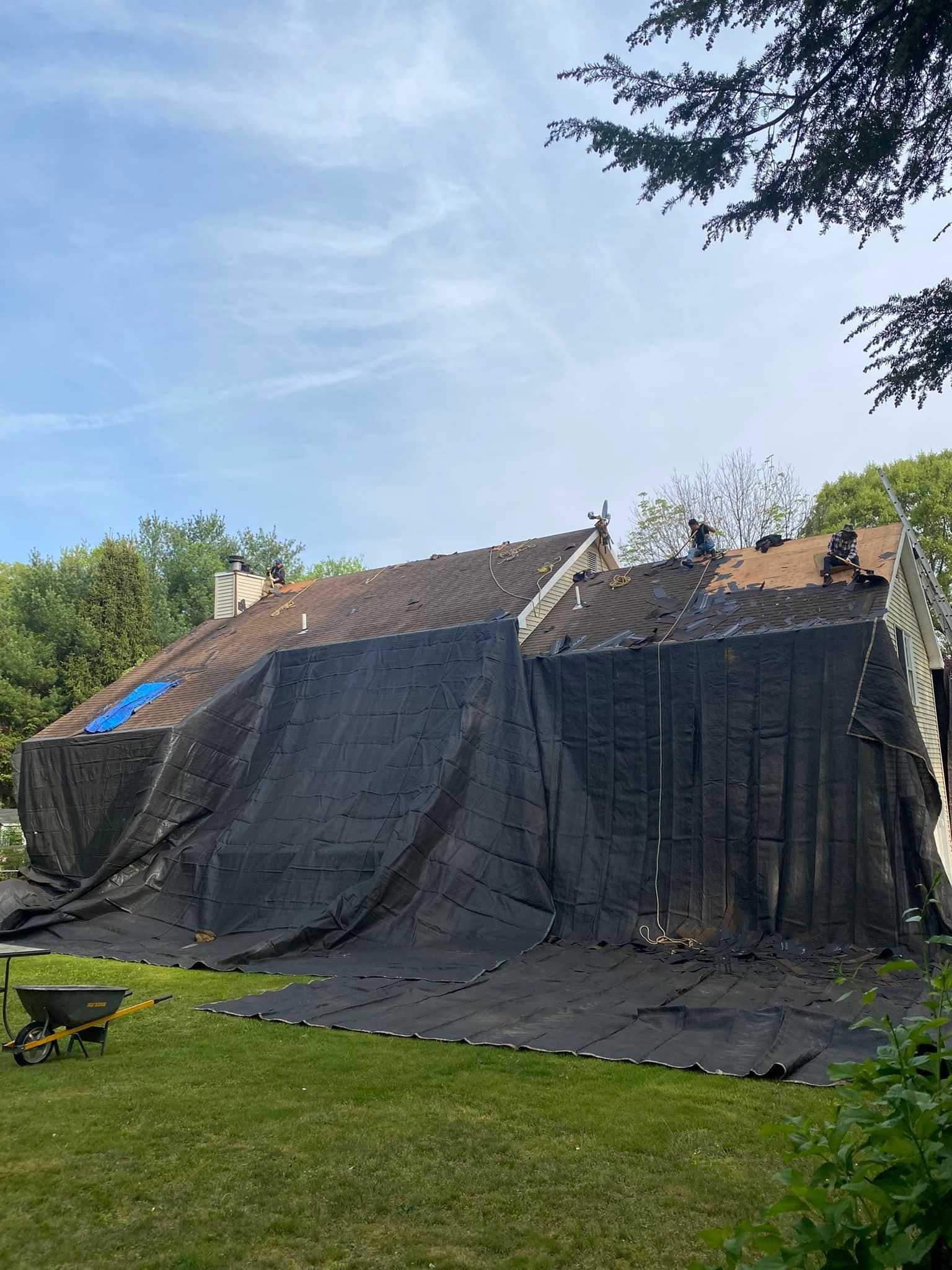 A house with a partially removed roof, covered with black tarp. Green grass and trees surround the structure