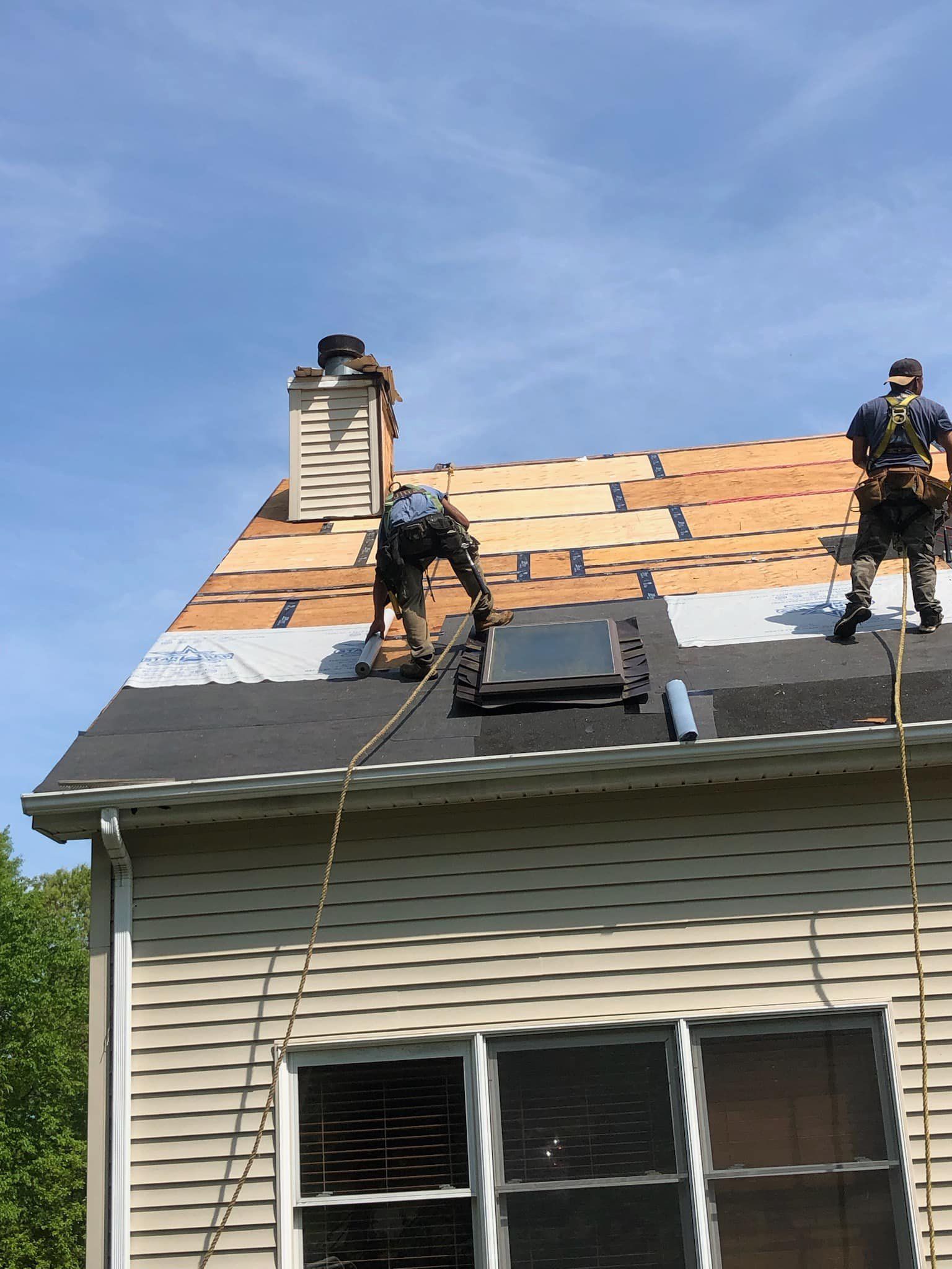 Two roofers installing new shingles on a house roof, secured by safety harnesses, with a chimney and skylight visible.