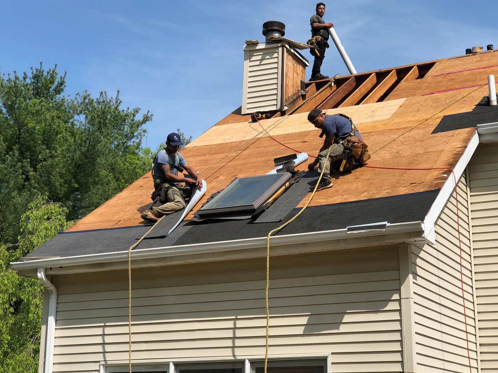 Three roofers working on a house roof, installing shingles and working near a chimney and skylight, on a sunny day.