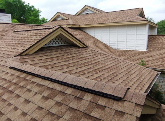 Brown shingle roof with dormers and a white exterior wall section; a tree line is visible in the background.
