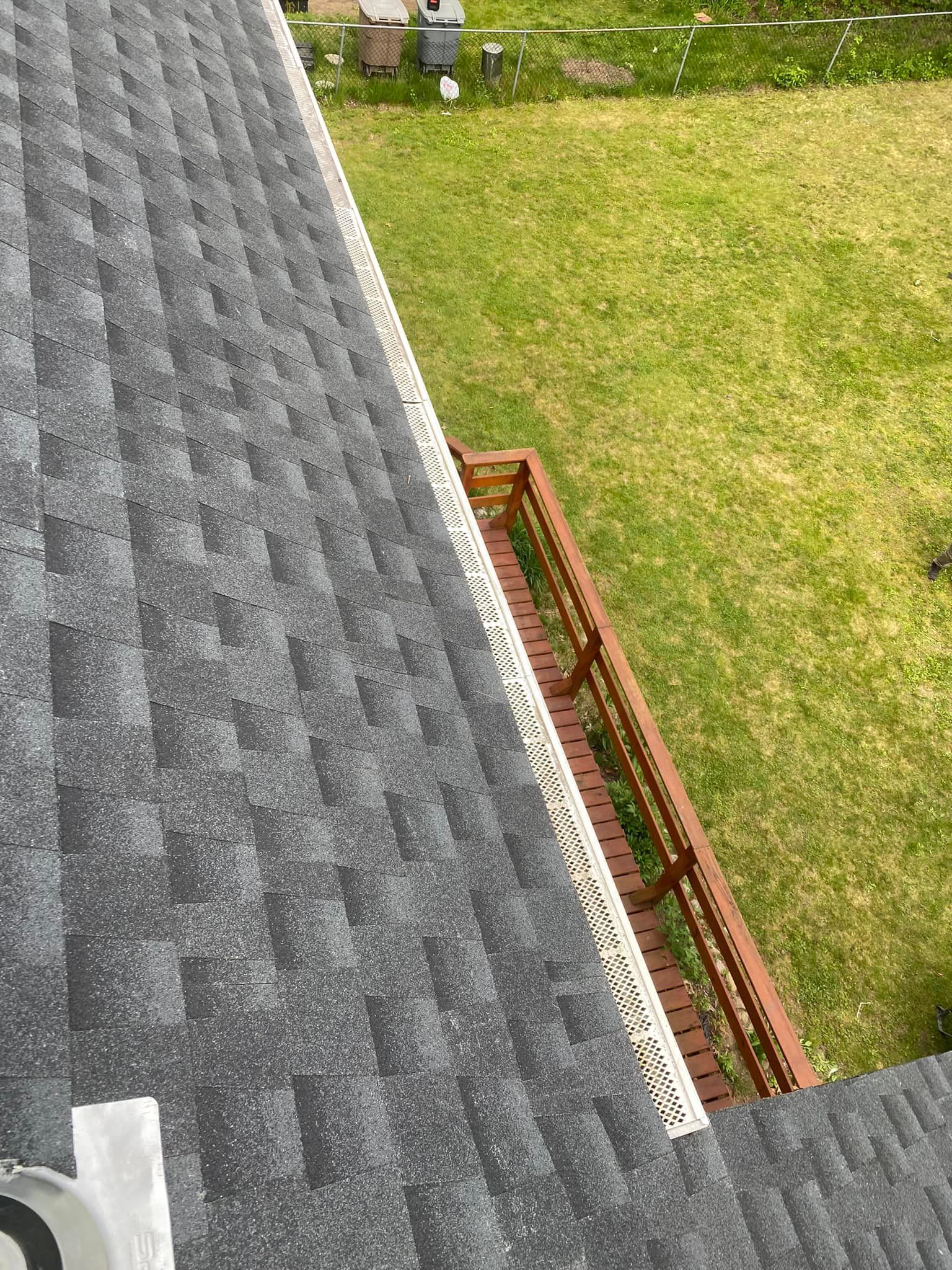 View from a rooftop overlooking a grassy yard and a wooden deck with white lattice. Gray shingles line the roof.