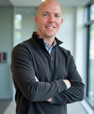 A man in a black quarter-zip sweater over a collared shirt smiling with arms crossed in a brightly lit office corridor.