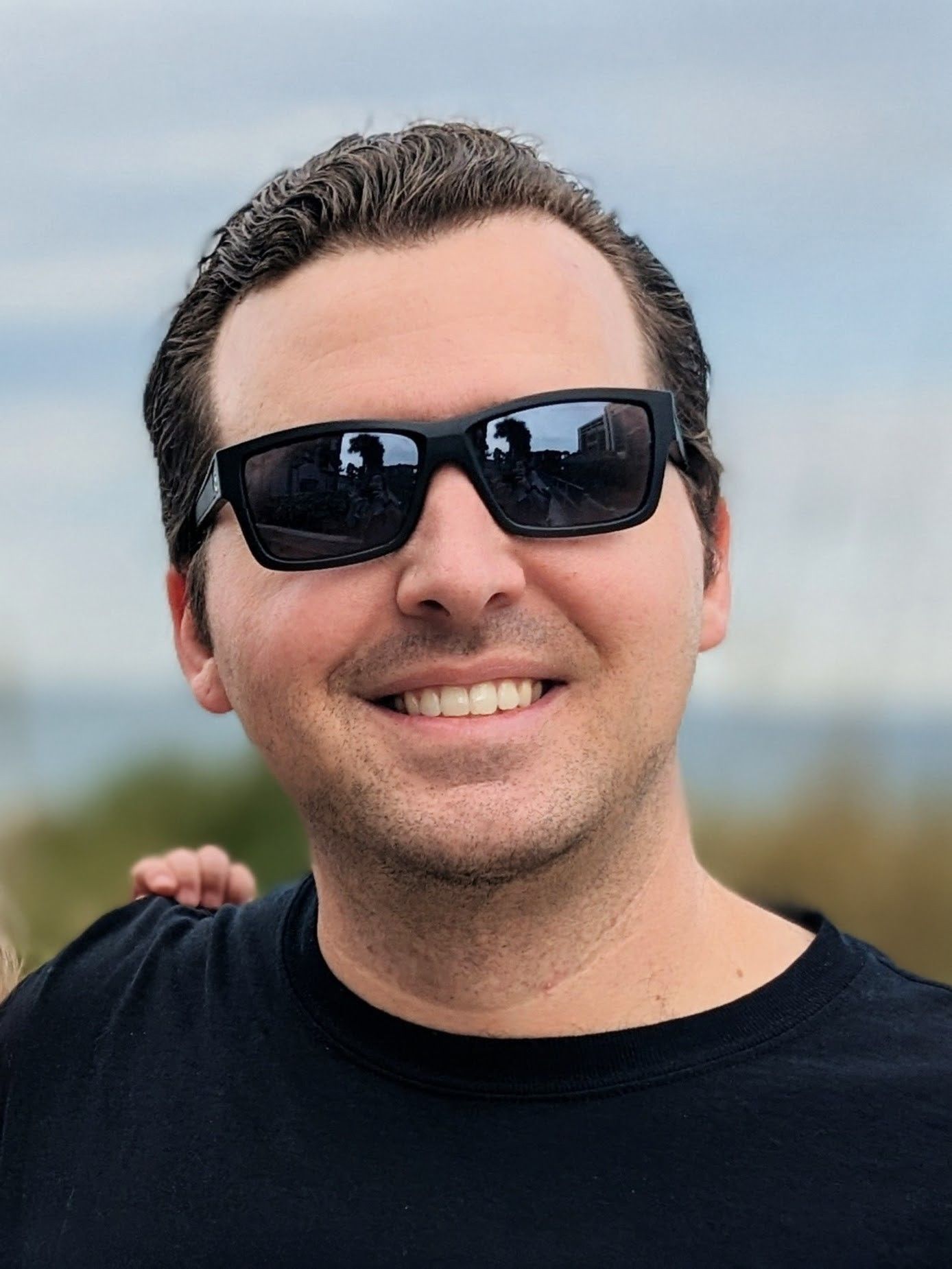 Man wearing black sunglasses smiles at the camera. Blue sky and water in background.