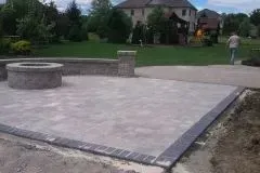 A man is walking down a sidewalk next to a fire pit in a backyard.