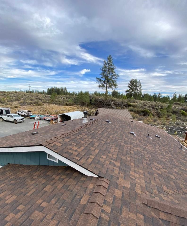 Brown shingled roof under a cloudy sky. Trucks and a tree are in the background.